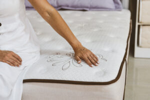 Close-up of a hand testing the memory foam layer of a white mattress, demonstrating the contouring effect and highlighting the detailed texture of the mattress cover in a bedroom setting.