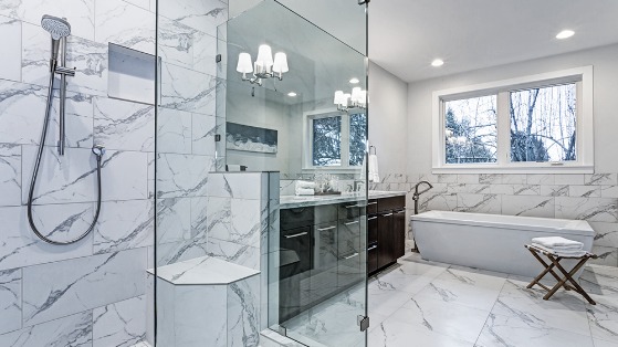 Spacious modern bathroom with marble tiles, glass shower enclosure, double vanity sink, and a freestanding bathtub near a window with a view of bare trees.