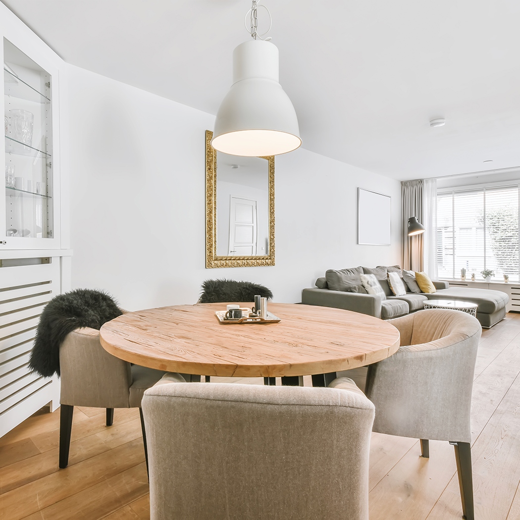 Modern dining area featuring a round wooden table surrounded by plush gray chairs, highlighted by a contemporary white pendant light. Adjacent to the dining area is a cozy living space with a gray sofa, paired with a large ornate gold-framed mirror on the wall. The room is illuminated by natural light from the window and exudes a calm, neutral palette.