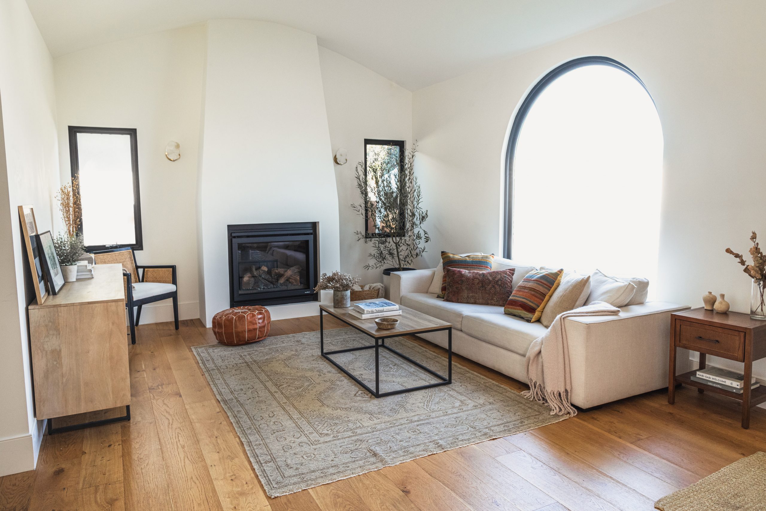 Minimalistic living room with a high arched window, neutral-toned sofa adorned with colorful throw pillows, built-in fireplace, wooden furniture, and a vintage-patterned area rug on hardwood flooring.