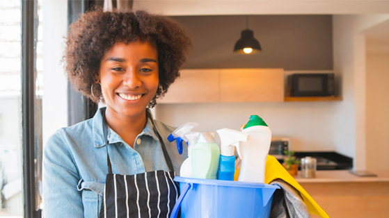 Smiling woman with curly hair holding a bucket of cleaning supplies in a modern kitchen setting.