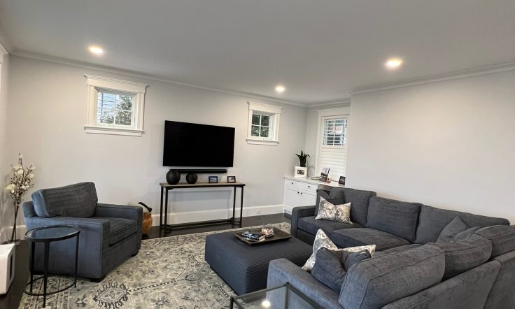 Modern living room with a gray sectional sofa, patterned rug, wall-mounted flat-screen TV, glass coffee table, and bright recessed lighting.