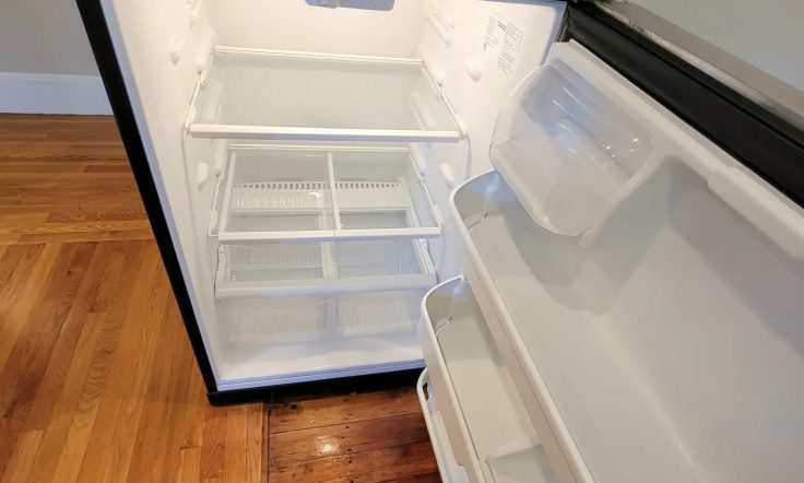 Open stainless steel refrigerator with empty shelves and door compartments, placed on wooden flooring in a room with bright lighting.