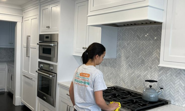 Cleaning professional in a white branded t-shirt wiping down a modern gas stove in a kitchen with white cabinets and a herringbone backsplash.