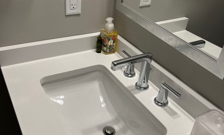 Modern bathroom with undermount sink, silver faucet, and clear soap dispenser on a white countertop, with electrical outlets and framed mirror above.