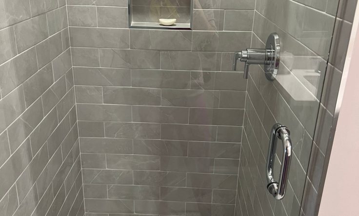 A modern shower stall with gray rectangular tiles on the walls. The shower features a built-in niche shelf for storing products, with a soap bar visible in the bottom section. The floor of the shower has smaller mosaic-style tiles with a central drain. On the right side, there's a silver showerhead and a matching handle, with a chrome grab bar positioned below.