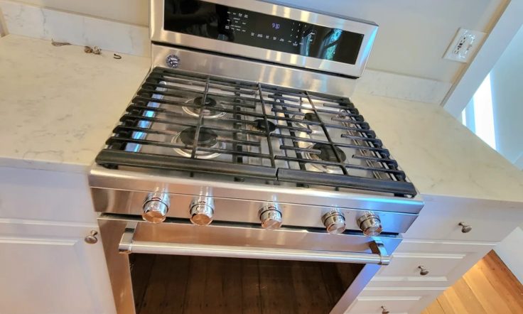 Stainless steel gas range with multiple burners, a digital display panel, and a matching oven below. The range is set into a white cabinet with a marble countertop, and the floor beneath is made of varnished wooden planks.