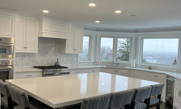 Modern kitchen interior with a spacious white marble island, upholstered bar stools, elegant cabinetry, and a herringbone backsplash, with a view of the cityscape through the windows.