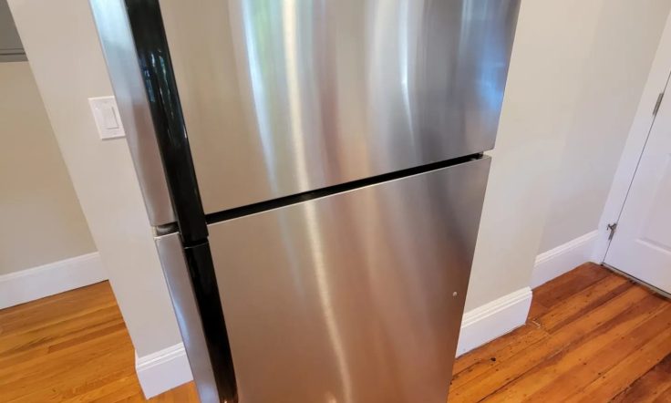 Stainless steel refrigerator with a top freezer and a bottom fridge section, positioned on wooden flooring next to a white wall.