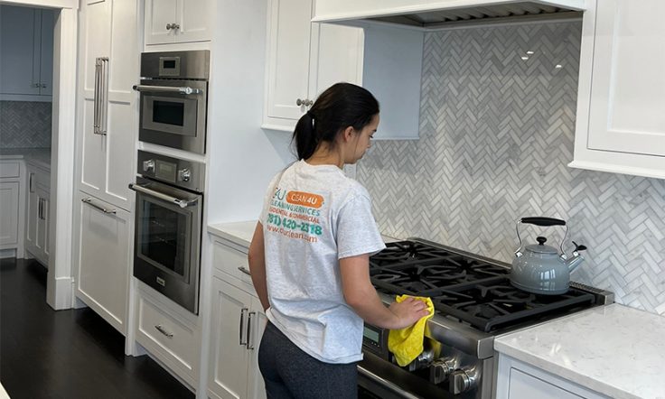 Cleaning professional in a white branded t-shirt wiping down a modern gas stove in a kitchen with white cabinets and a herringbone backsplash.