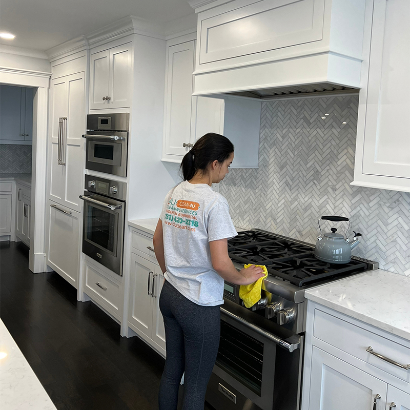 Cleaning professional in a white branded t-shirt wiping down a modern gas stove in a kitchen with white cabinets and a herringbone backsplash.