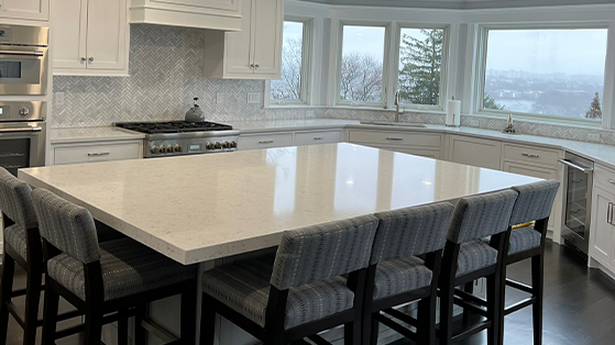 Modern kitchen interior with a spacious white marble island, upholstered bar stools, elegant cabinetry, and a herringbone backsplash, with a view of the cityscape through the windows.