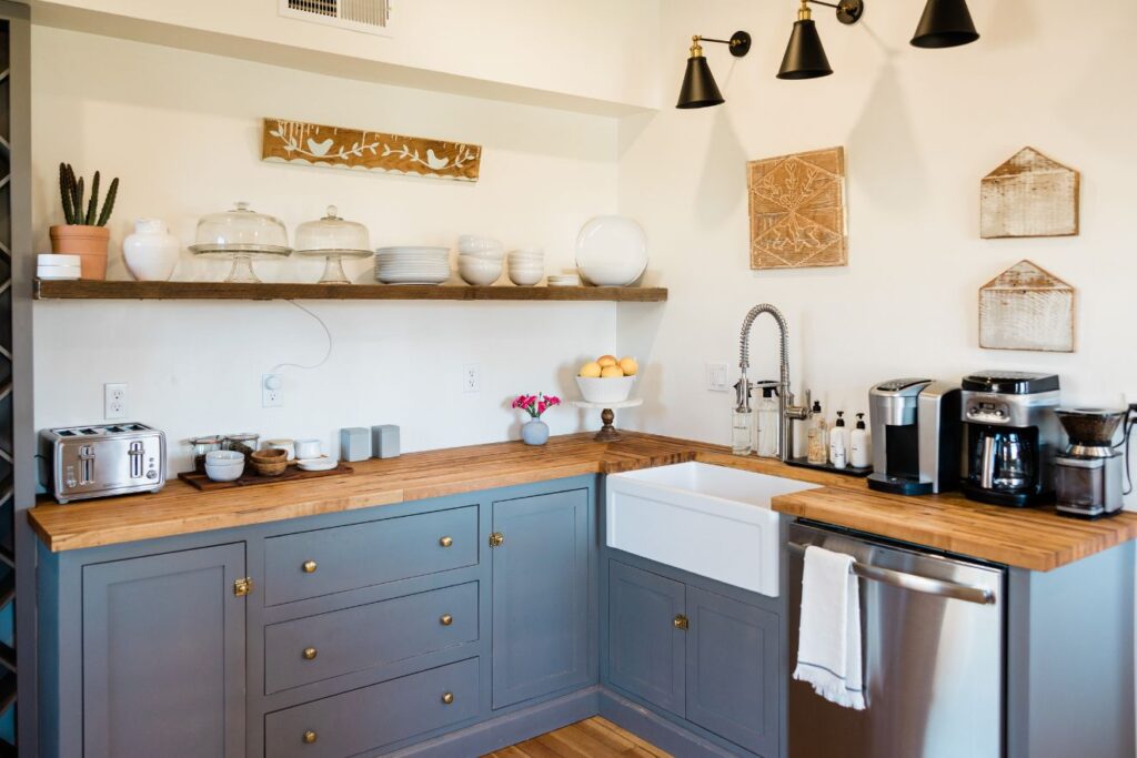 Rustic kitchen setup featuring wooden countertops, blue cabinetry with gold handles, wall-mounted wooden shelves displaying a variety of kitchenware, and an assortment of modern appliances. The decor is complemented by wall art, pendant lighting, and fresh produce and flowers adding a touch of warmth and homeliness.