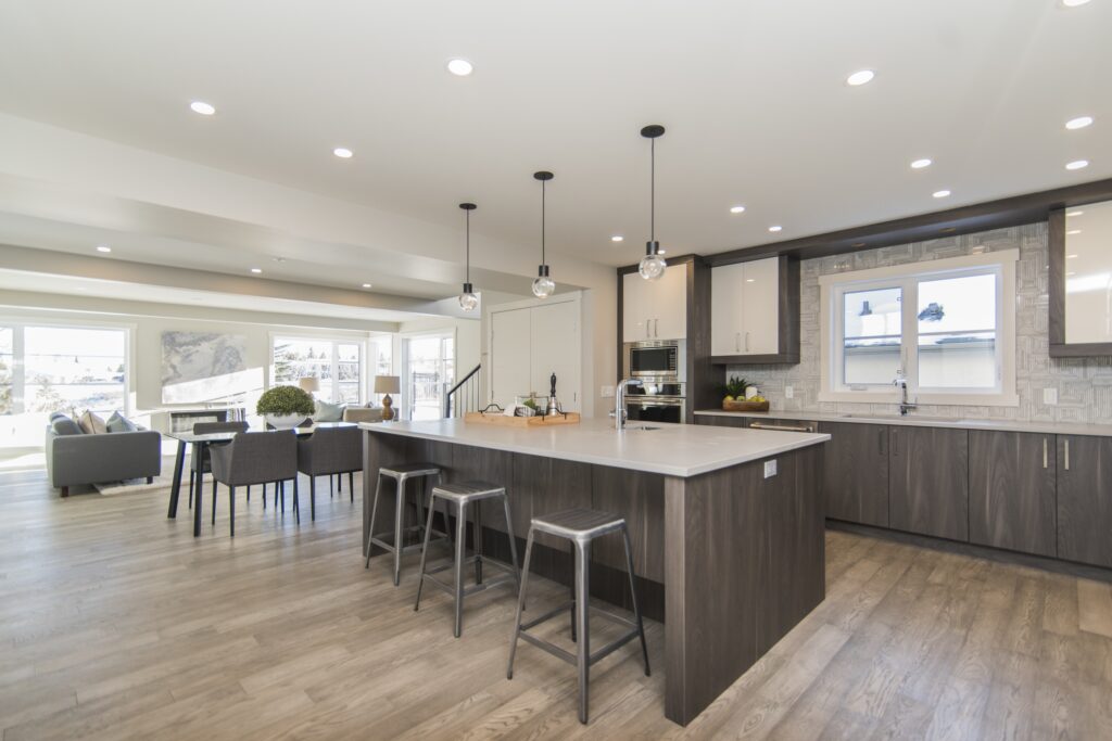 Elegant and contemporary open-concept living space featuring a kitchen with a dark wood-finished island and matching cabinets. The island is accompanied by modern bar stools and boasts a sleek white countertop. Pendant lights dangle above, providing illumination alongside recessed ceiling lights. Adjacently, a cozy sitting area is set up with a gray sofa and complemented by framed artwork. Large windows around the space ensure plenty of natural light, enhancing the room's inviting and spacious feel.