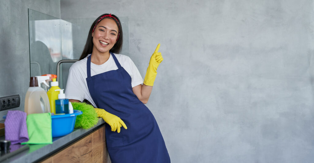 Woman in blue apron and yellow gloves smiling and pointing, surrounded by cleaning supplies in a modern kitchen setting.