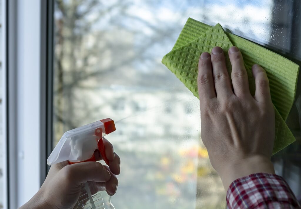 Hand holding a spray bottle and cleaning window glass with a green microfiber cloth, showing the removal process.