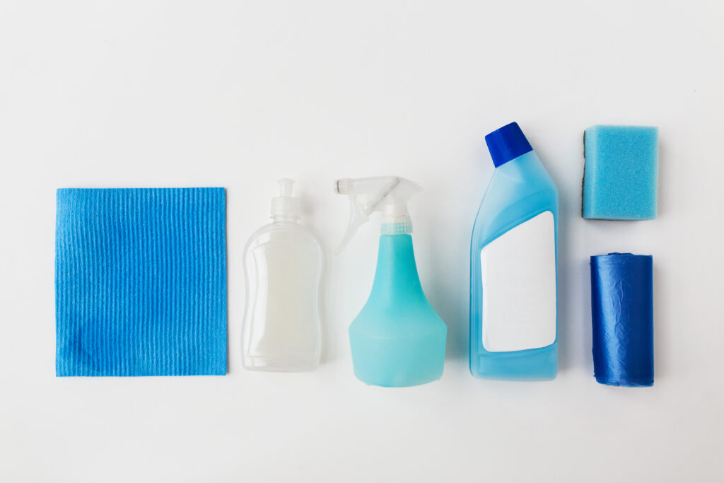 Blue and transparent cleaning products, including cloth, spray bottles and soap, sponge, and trash bag, arranged on a white background