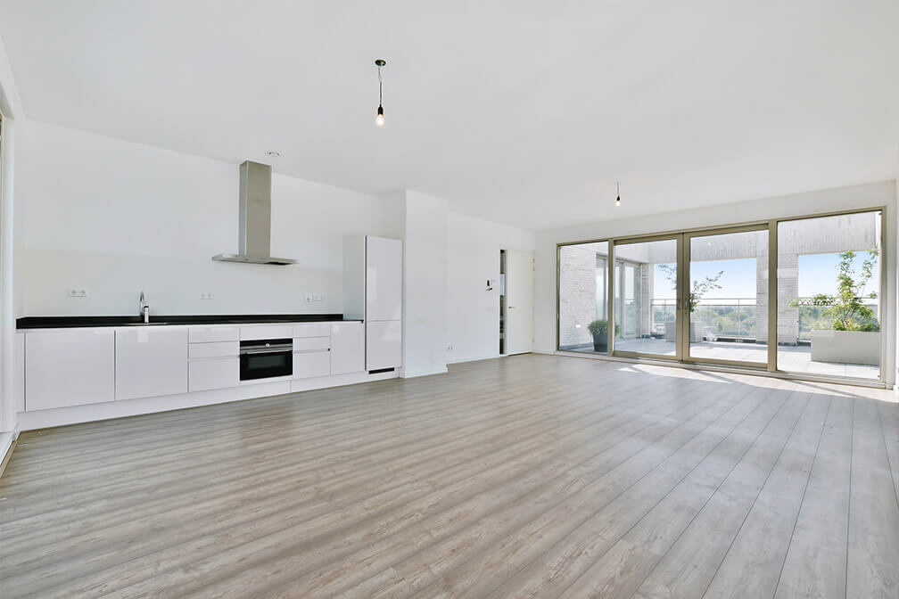 Spacious, minimalist kitchen and living area featuring a white modern kitchen setup with a built-in oven, sleek exhaust hood, and a black countertop. The wooden flooring complements the pristine ambiance. Large sliding glass doors lead to a balcony, allowing for an abundance of natural light and offering views of the outside.