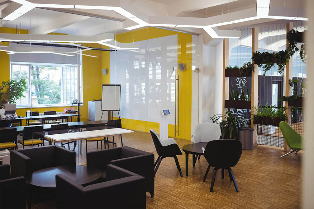 Modern co-working space with vibrant yellow accent walls, a variety of seating options from sofas to chairs, and an array of plants. The area is illuminated by sleek overhead lights and features large windows offering ample daylight.