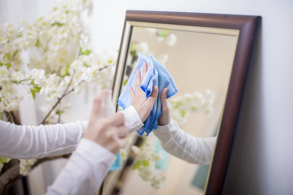 Hands cleaning a framed mirror with a blue cloth, with white blossoming flowers in the background.